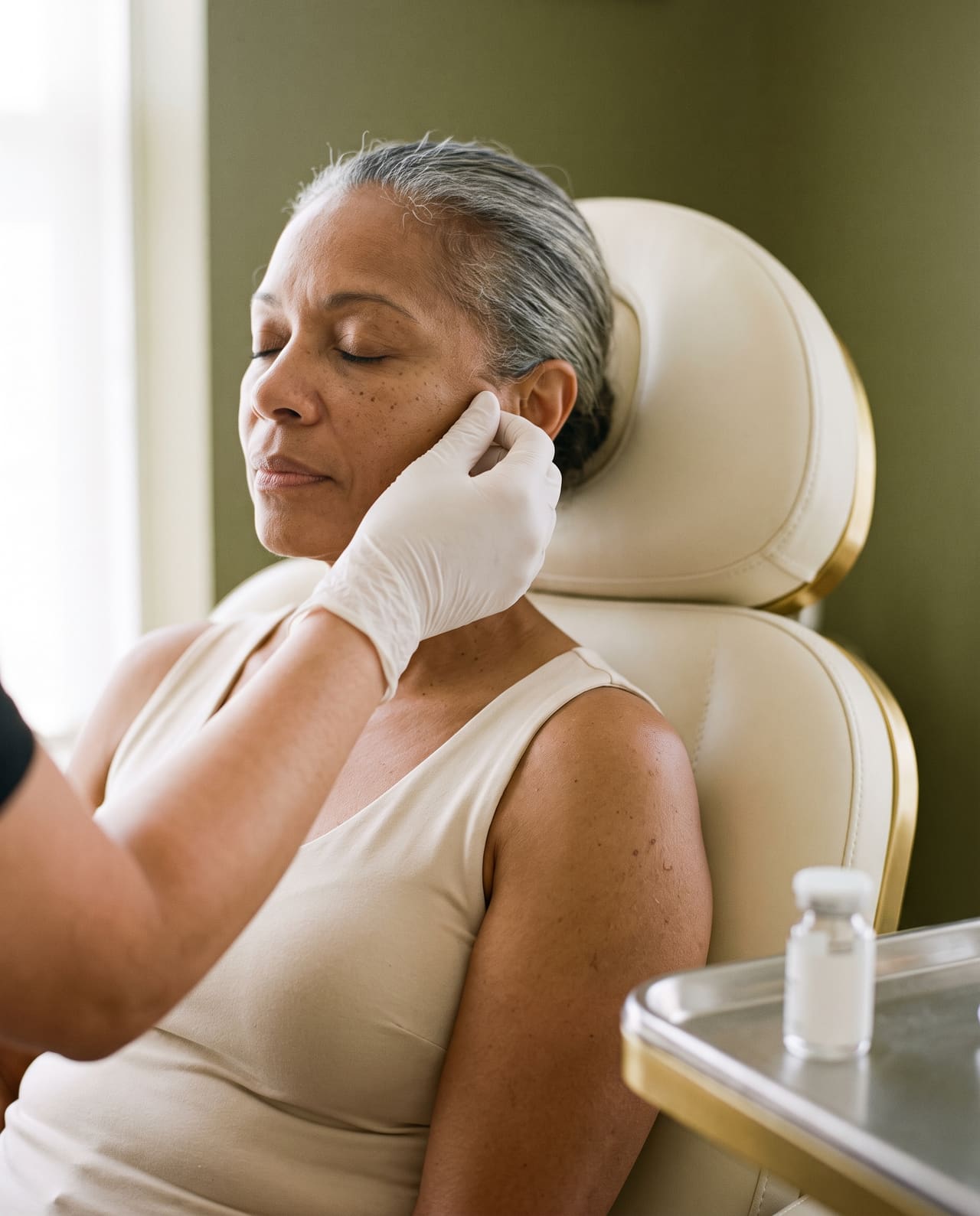 A gloved hand resting on the cheek of a mature client with silver hair, a small unlabeled vial on the tray beside her.
