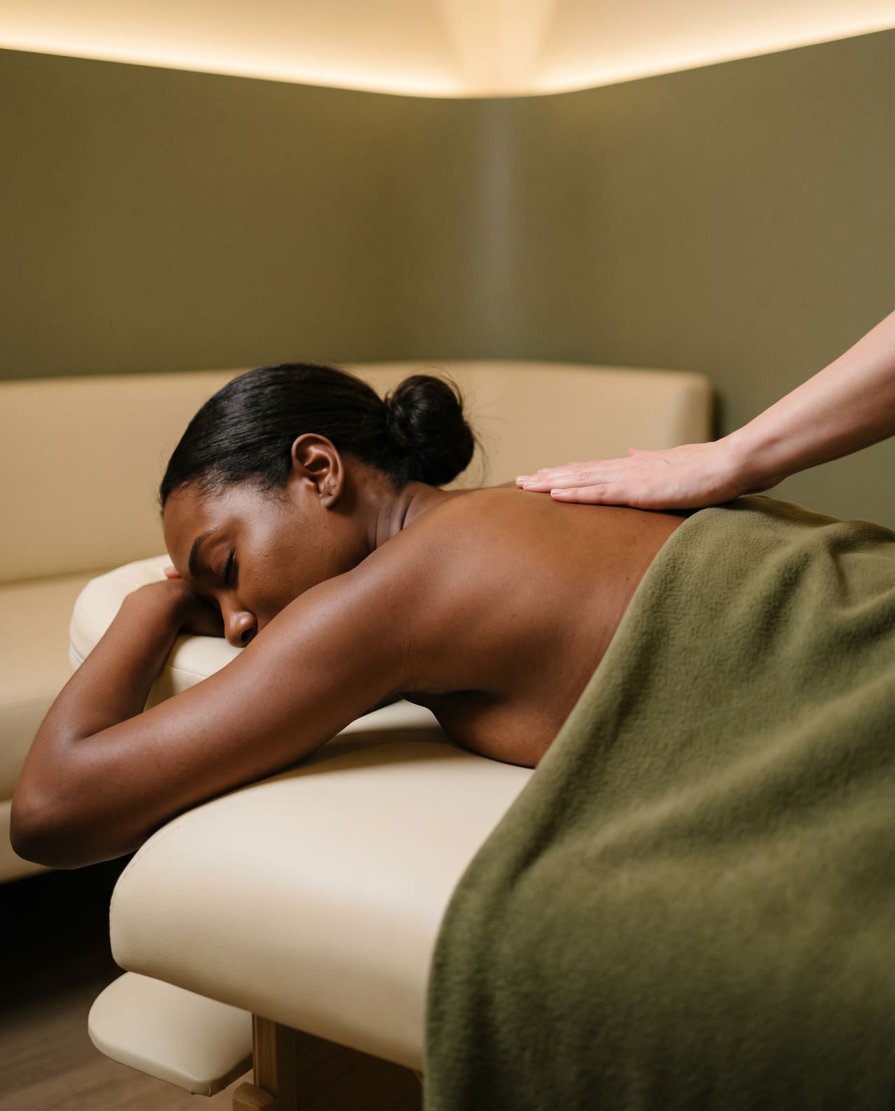 A hand resting on the upper back of a client lying face down on a massage bed under a green blanket.
