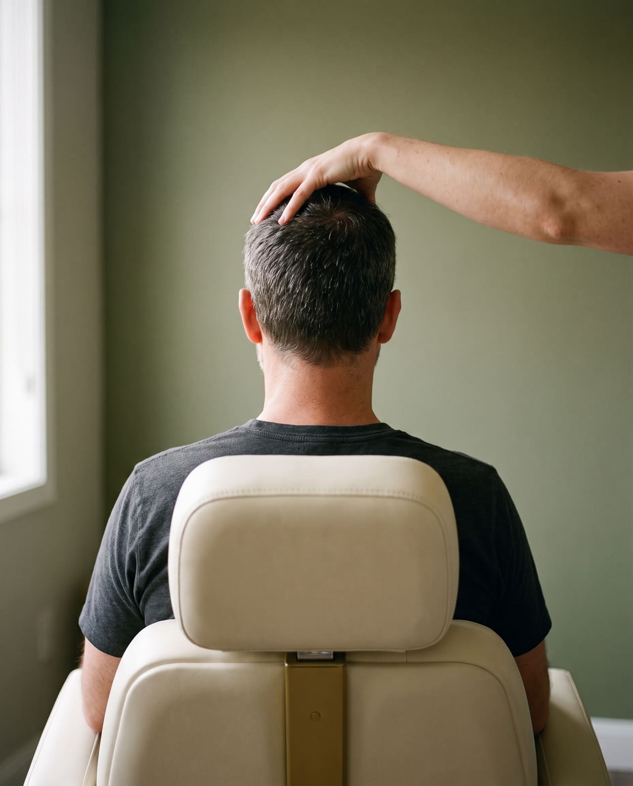 A hand resting on the crown of a seated man's head, seen from behind, during a scalp assessment.