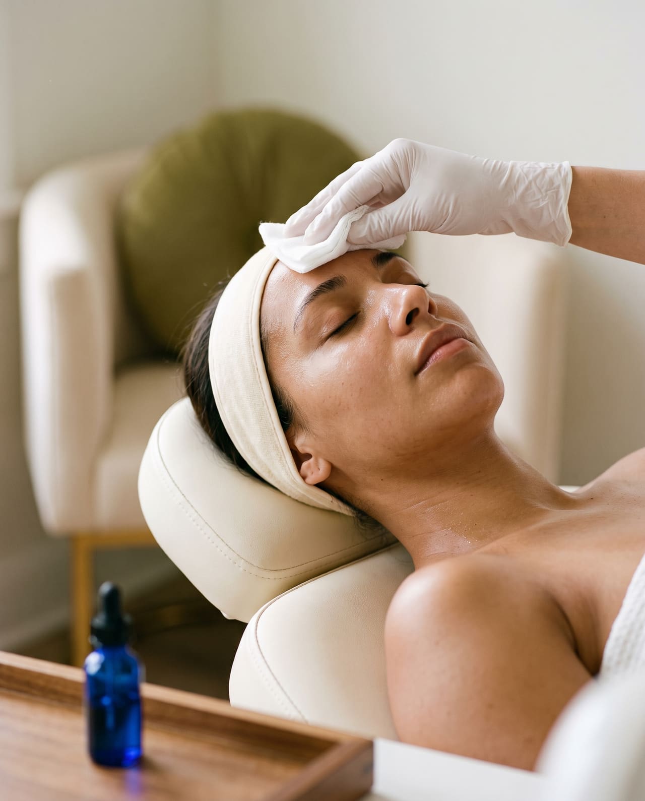 A gloved hand holding a cotton gauze against the forehead of a reclined client, a dropper bottle on the counter.